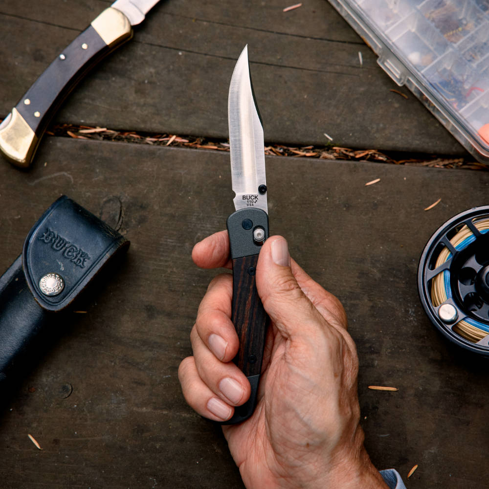 Buck 110 Folding Hunter Crossbar Knife in Hand with Traditional Buck 110 in Background